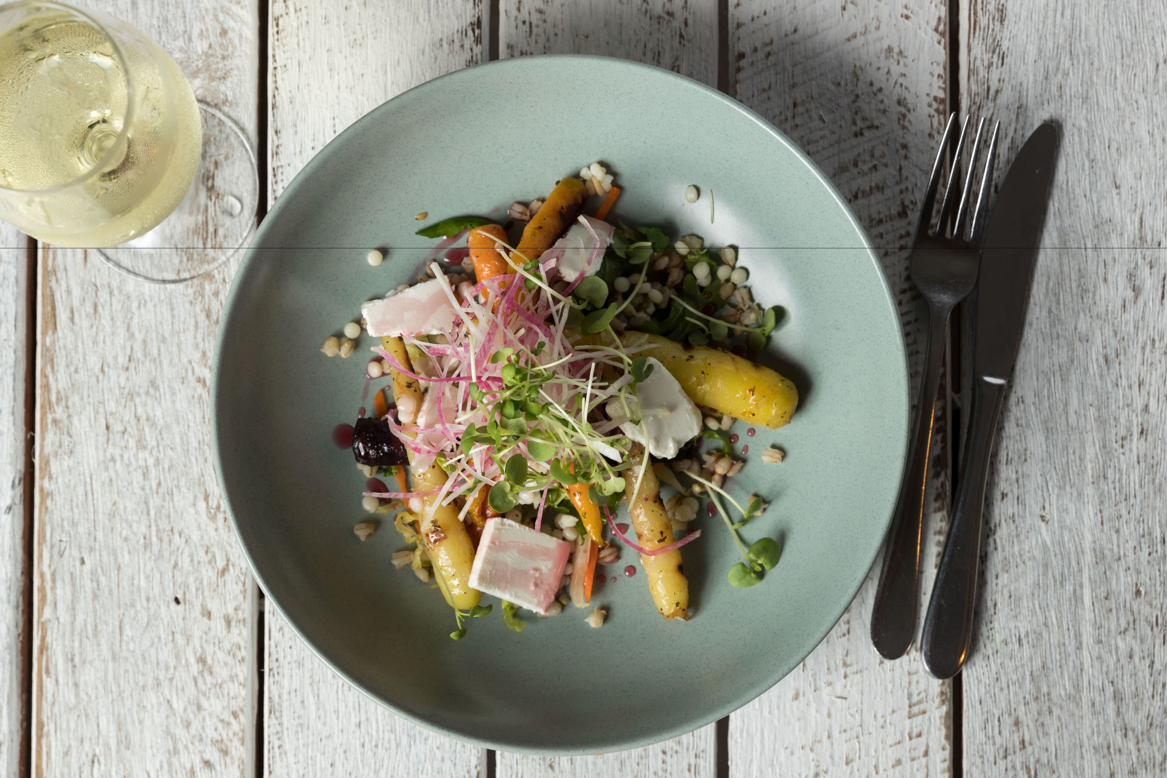 A plate of food with various vegetables and grains, accompanied by a glass of white wine, on a rustic wooden table.