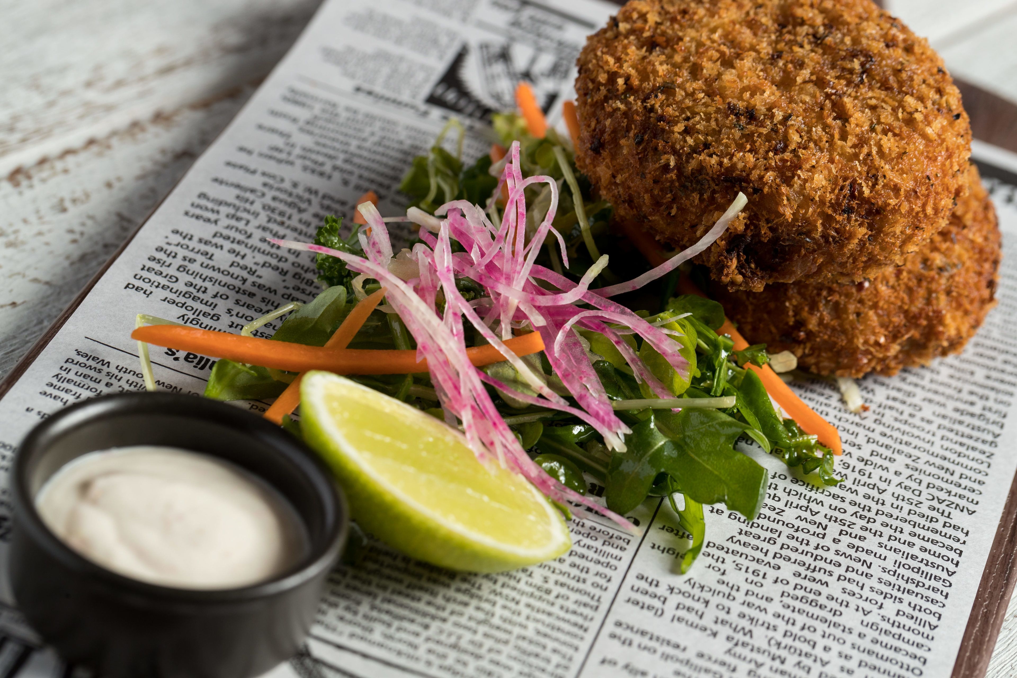 A meal with a breaded item, salad, and a small bowl of sauce on a newspaper.