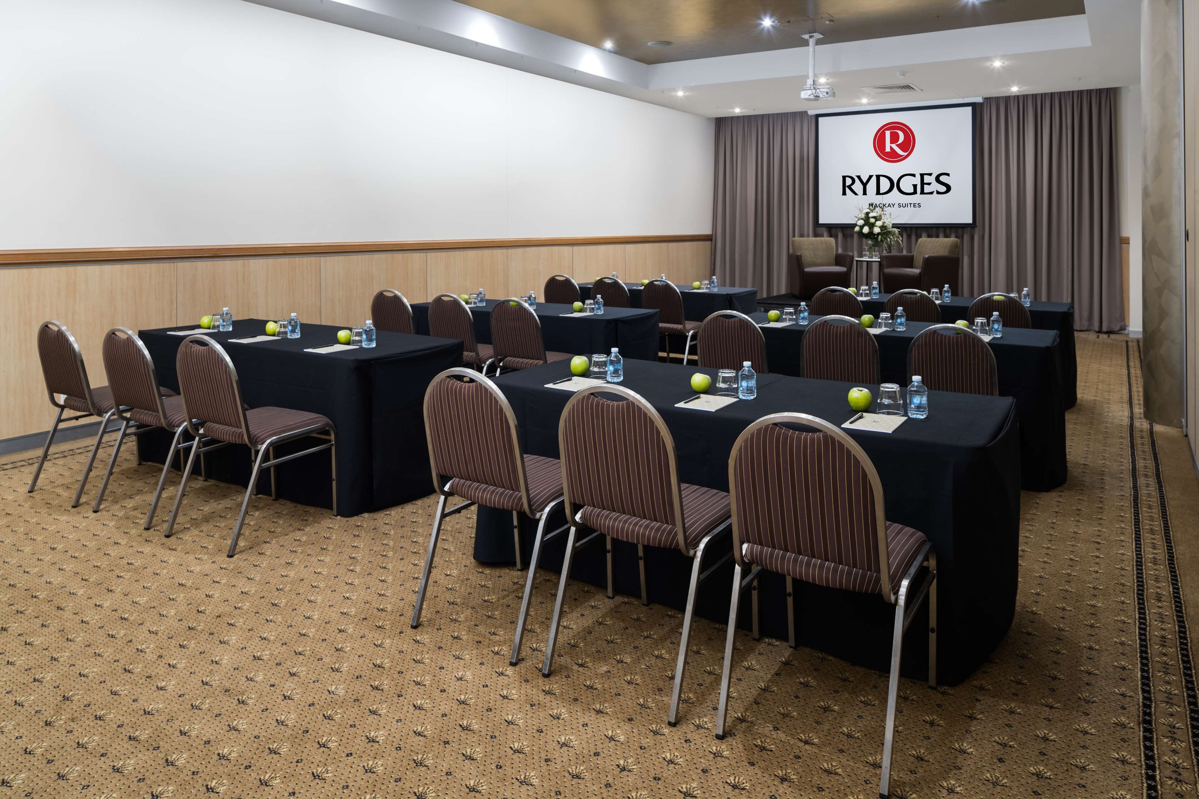 A conference room with multiple tables and chairs, a projector screen displaying the Rydges logo, and a carpeted floor.