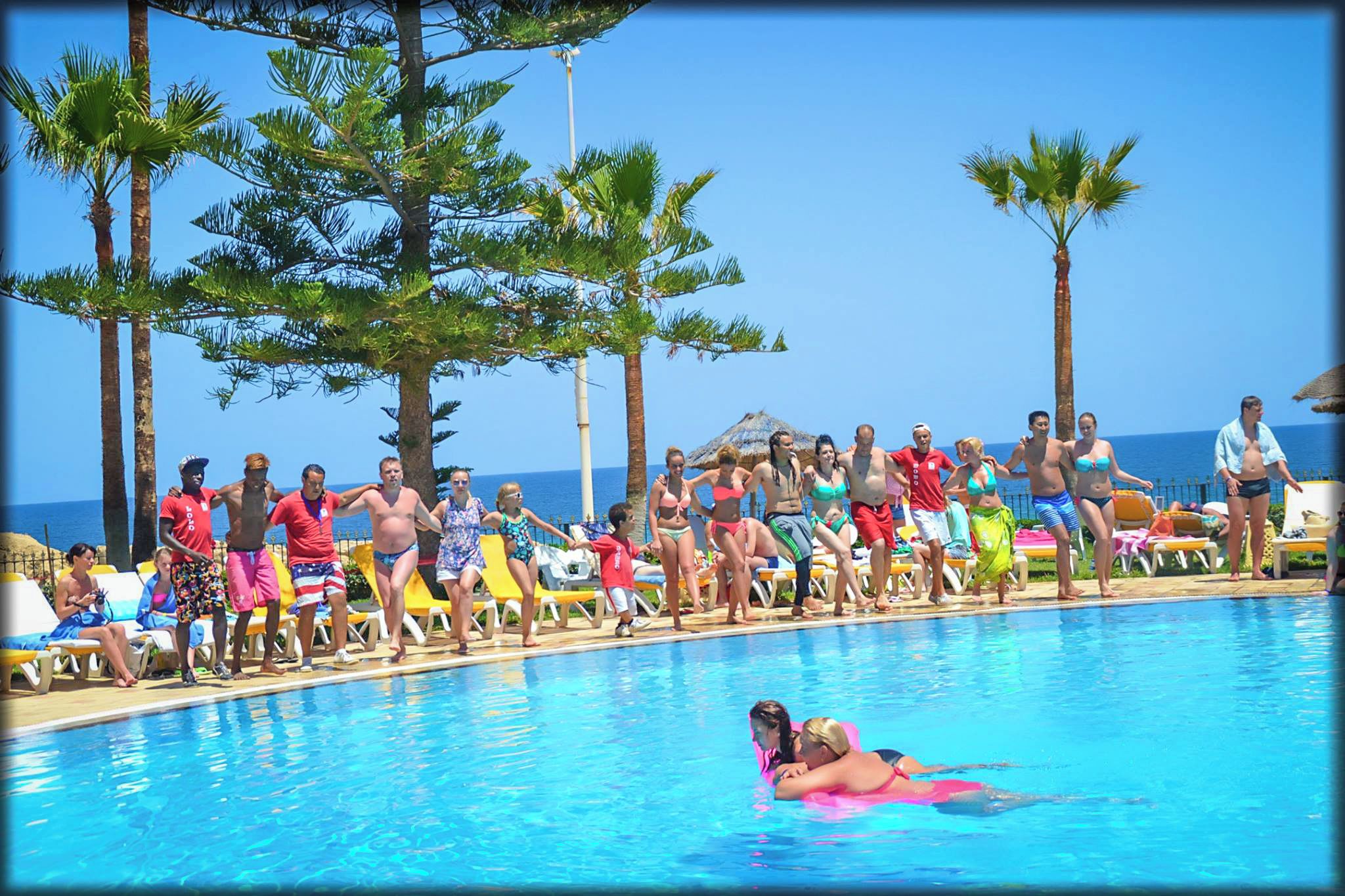 Zona junto a la piscina con gente nadando y tomando el sol, rodeada de palmeras y una vista despejada del océano.
