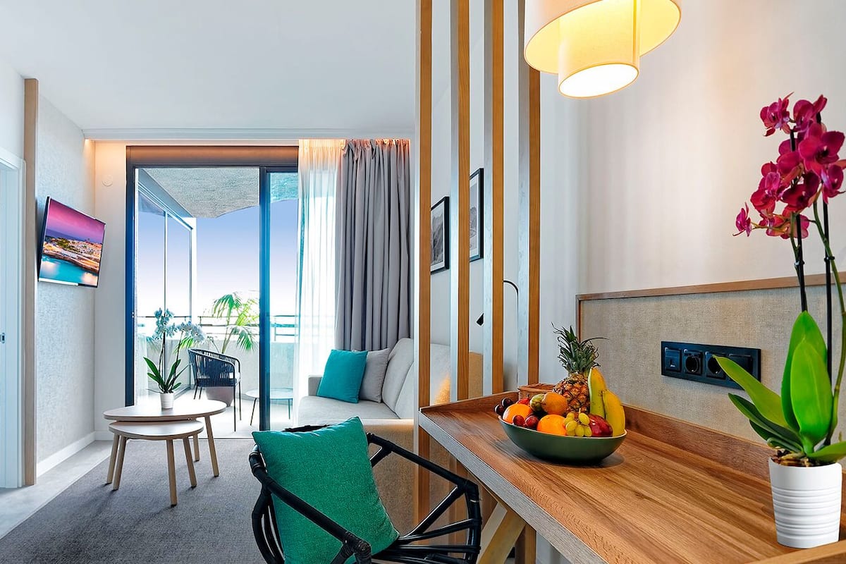 A modern hotel room with a dining area, a view of the outdoors, and a fruit bowl on the table.