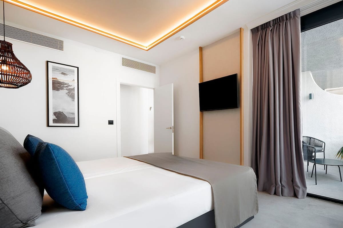 A modern hotel room with a bed, a TV, a balcony with a chair and a plant, and a large window.