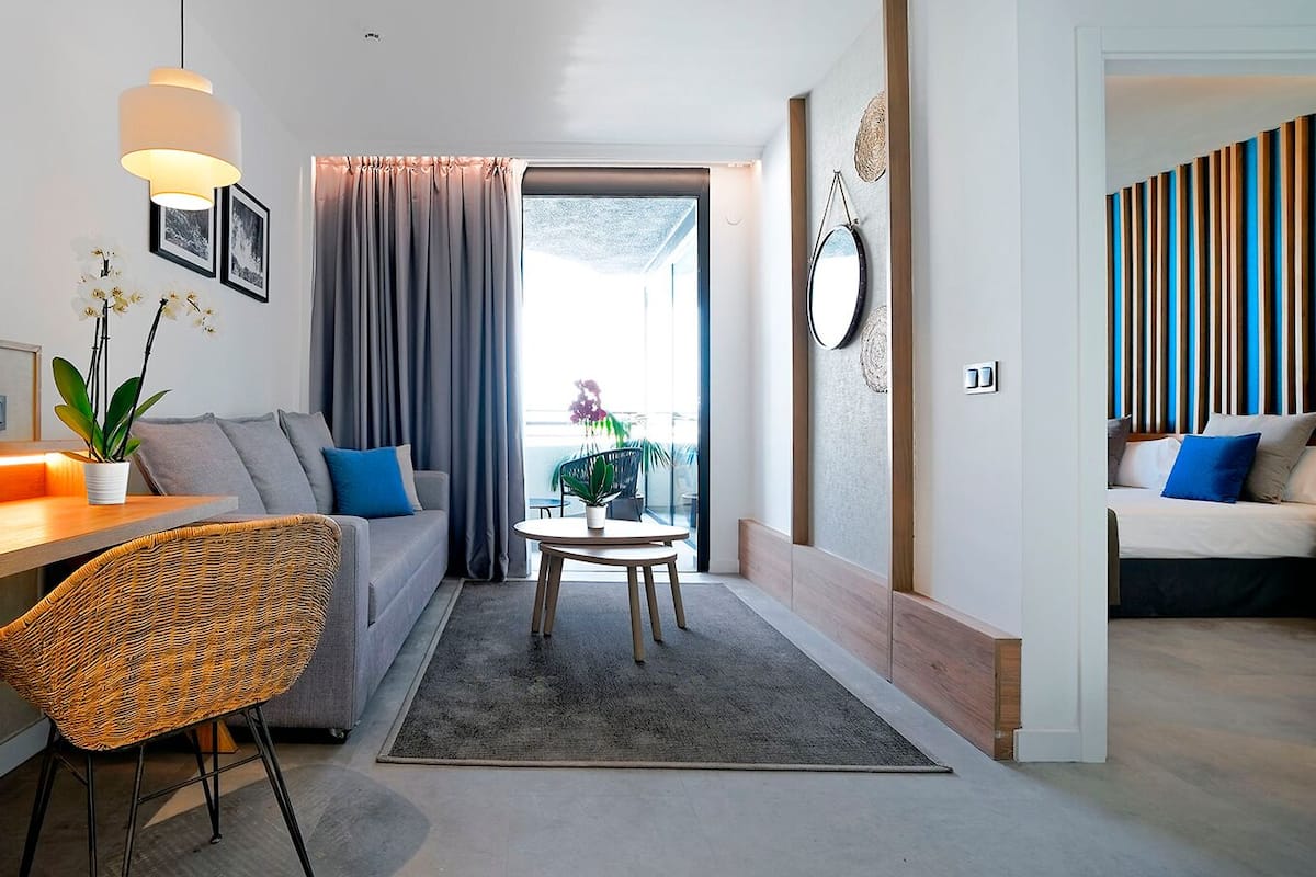 A modern living room with a sofa, a dining area with a table and chairs, and a view through a large window.
