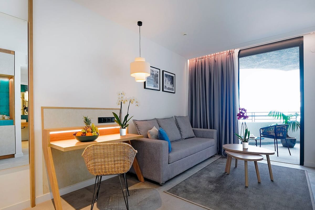 A modern living room with a sofa, a dining table, a small kitchen area, and a balcony with a view.