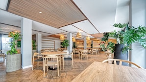 Un comedor moderno con mesas y sillas de madera, plantas grandes en macetas y techo de madera.