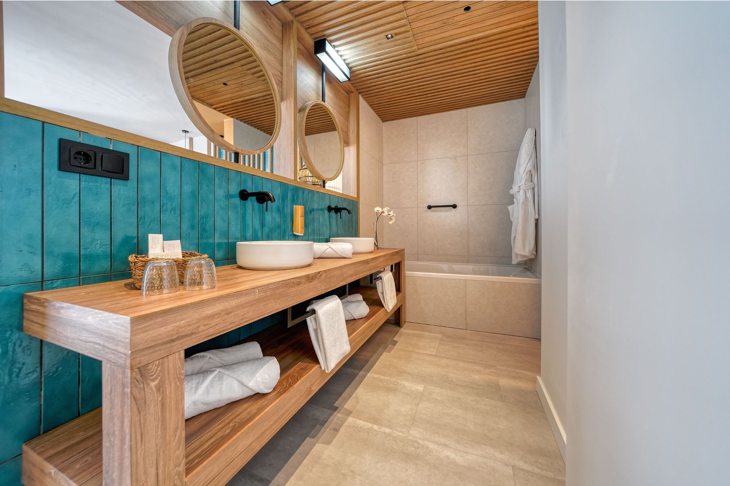 A modern bathroom with wooden vanity, two large mirrors, a bathtub, and a teal wall.
