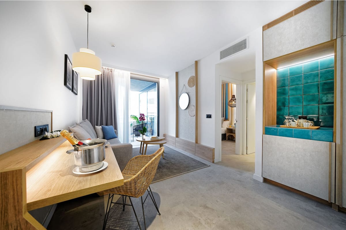 A modern hotel room with a wooden dining table, wicker chairs, a minibar, and a view of the outdoors.