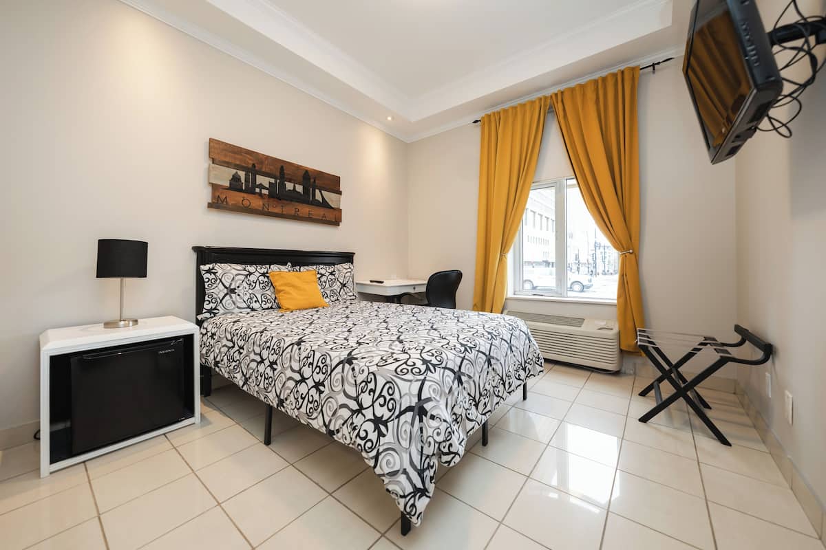 Quarto com cama, escrivaninha, cadeira, televisão e janela com cortinas.