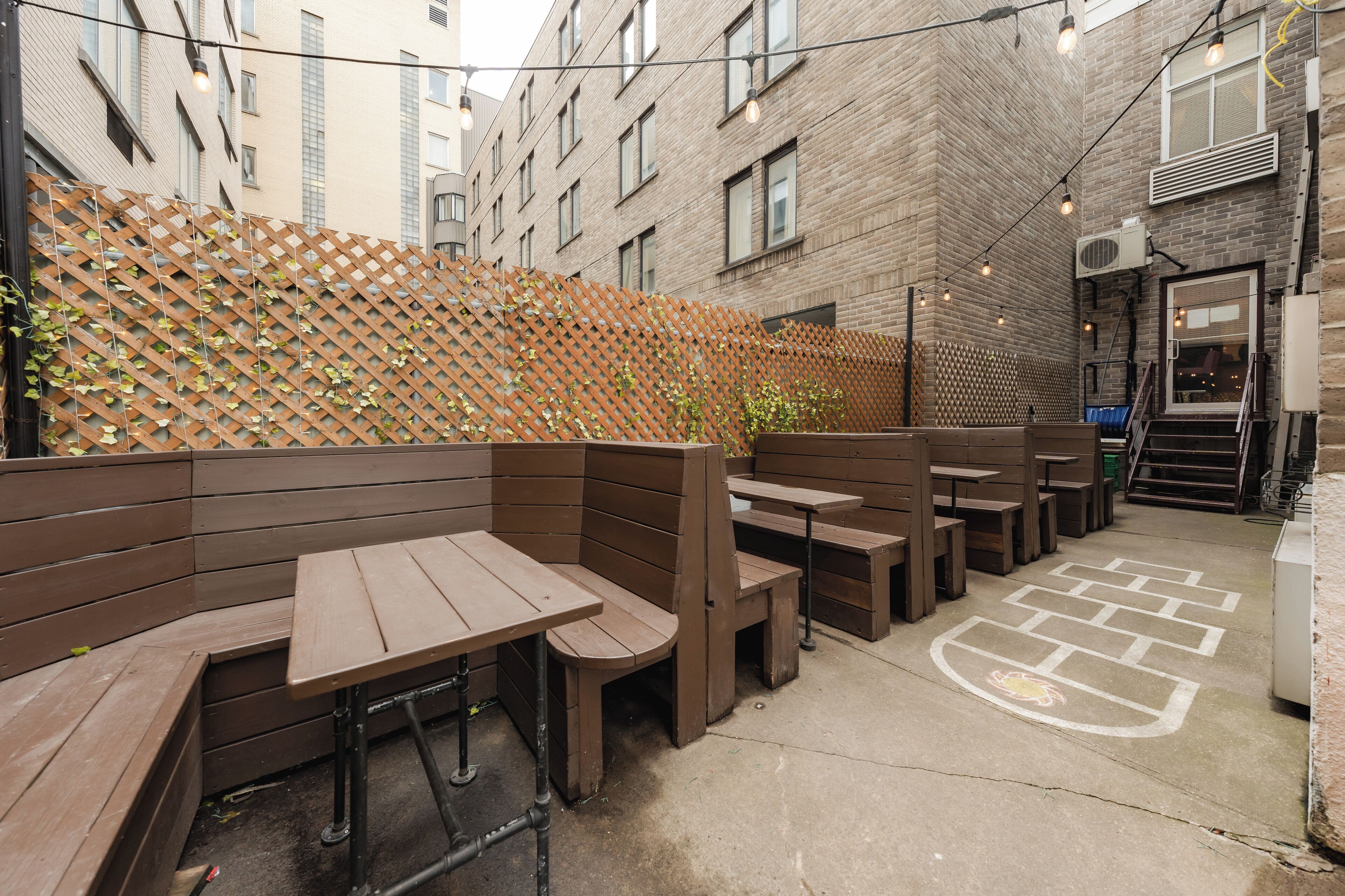 An outdoor seating area with wooden benches and tables, a brick wall with climbing plants, and a concrete floor with a chalk drawing.