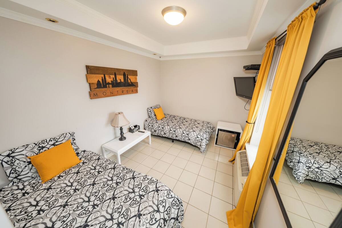 Quarto com duas camas, uma mesa pequena, televisão e janela com cortinas amarelas.