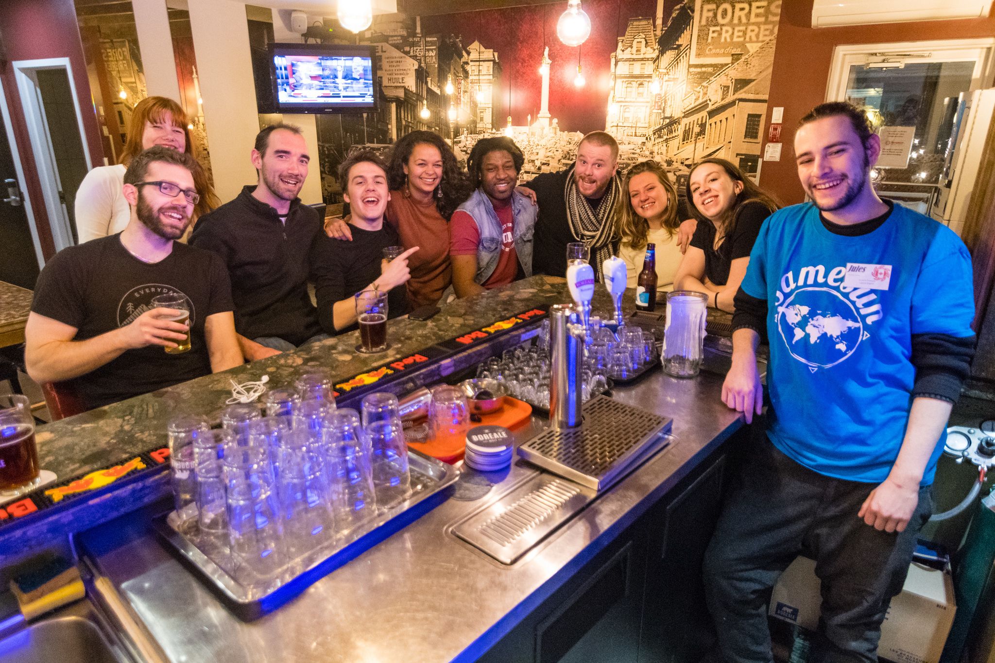 Un groupe de personnes est réuni dans un bar, avec un comptoir, des verres et un téléviseur en arrière-plan.