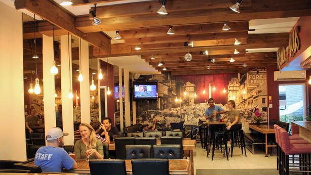Un restaurant avec des poutres en bois, des luminaires suspendus, des tables et des chaises, et un écran de télévision diffusant un programme d’information.