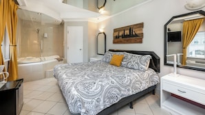 Une chambre avec un grand lit, une table de chevet, un miroir, une œuvre d’art représentant un paysage urbain et un espace douche.