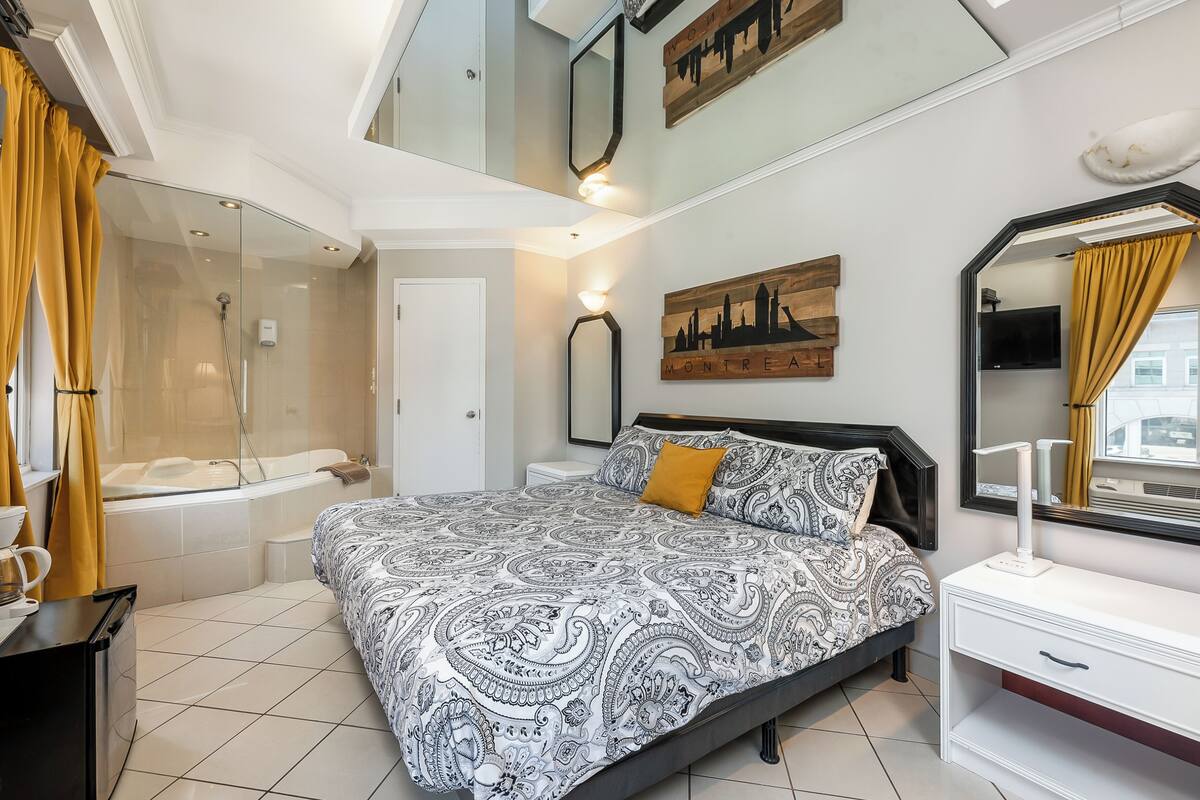 Quarto com cama grande, mesa de cabeceira, espelho, uma obra de arte com vista para a cidade e área de chuveiro.