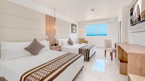 A hotel room with two beds, a desk, and a view of the sea.