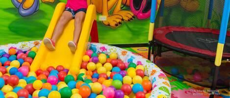 A child on a yellow slide in a ball pit with colorful balls, surrounded by cartoon characters on the wall.