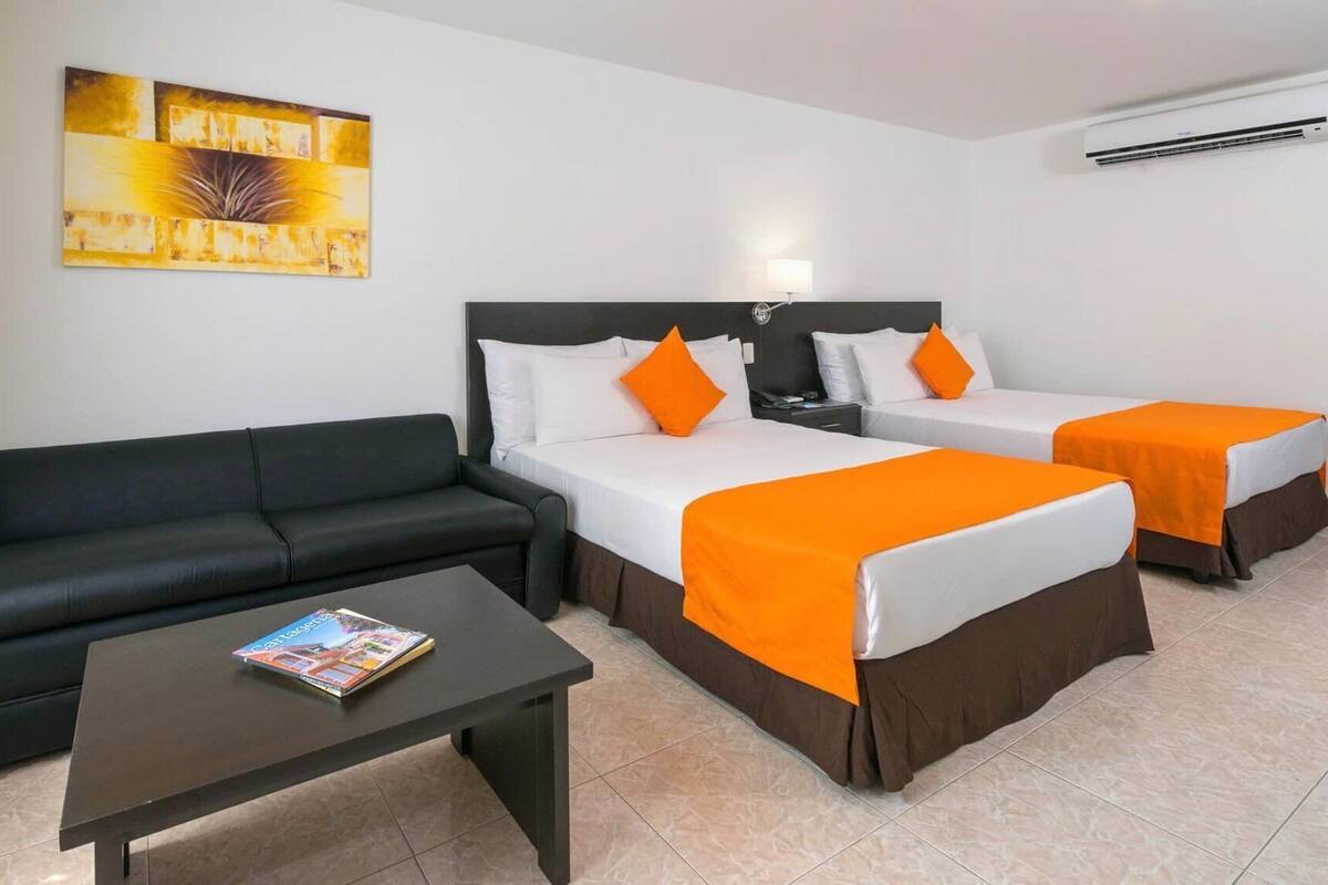 Een hotelkamer met een zwarte bank, twee eenpersoonsbedden met oranje beddengoed, een klein tafeltje met een tijdschrift, en een schilderij aan de muur.