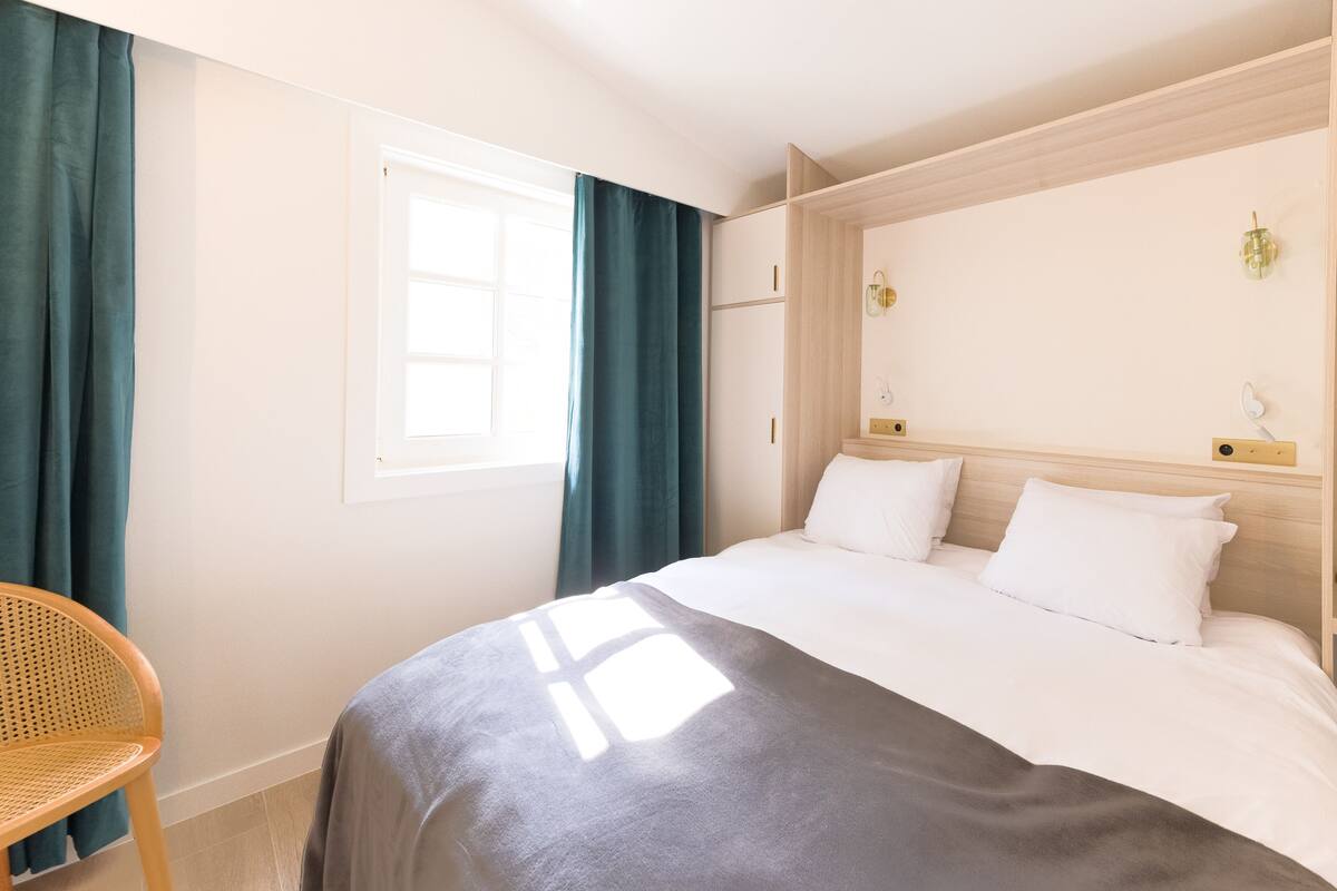 A hotel room with a bed, a chair, a window with curtains, and a wall-mounted light fixture.