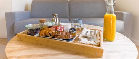 A breakfast tray with pastries, a bottle of orange juice, and a coffee pot on a round wooden table in a living room with a grey sofa.