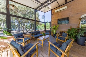 Un amplio espacio al aire libre con mobiliario de madera y grandes ventanales con vistas a una zona boscosa.