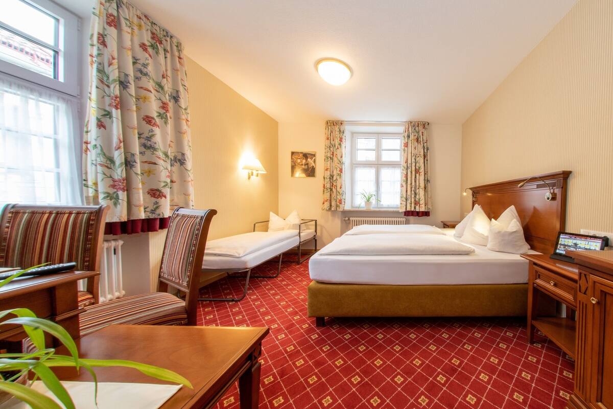 Ein Hotelzimmer mit Doppelbett, zwei Einzelbetten, einem Schreibtisch, einem Stuhl, einer Lampe und einem Vorhangfenster.