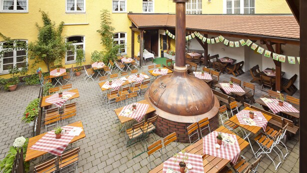 Une terrasse extérieure avec plusieurs tables et chaises en bois, un grand four en briques et un bâtiment jaune en arrière-plan.