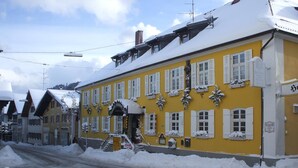 Un bâtiment jaune avec des volets blancs, de la neige sur le toit et les fenêtres, et une enseigne indiquant « Gasthof Gasthof ».