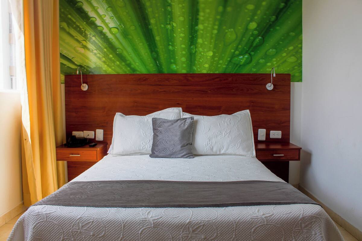 Ein Hotelzimmer mit einem hölzernen Kopfteil, einem Bett mit weißer Bettwäsche, einem grauen Kopfkissen und einem Nachttisch mit Telefon und Lampe.