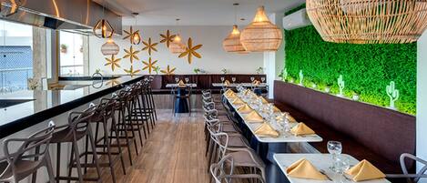 Um restaurante com um balcão comprido, várias mesas e cadeiras. Há luminárias pendentes e uma parede verde com plantas.