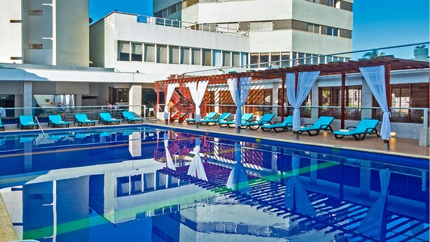 A swimming pool with blue lounge chairs and a covered area with white curtains. - Hotel Dorado Plaza Bocagrande - All Inclusive (Cartagena)