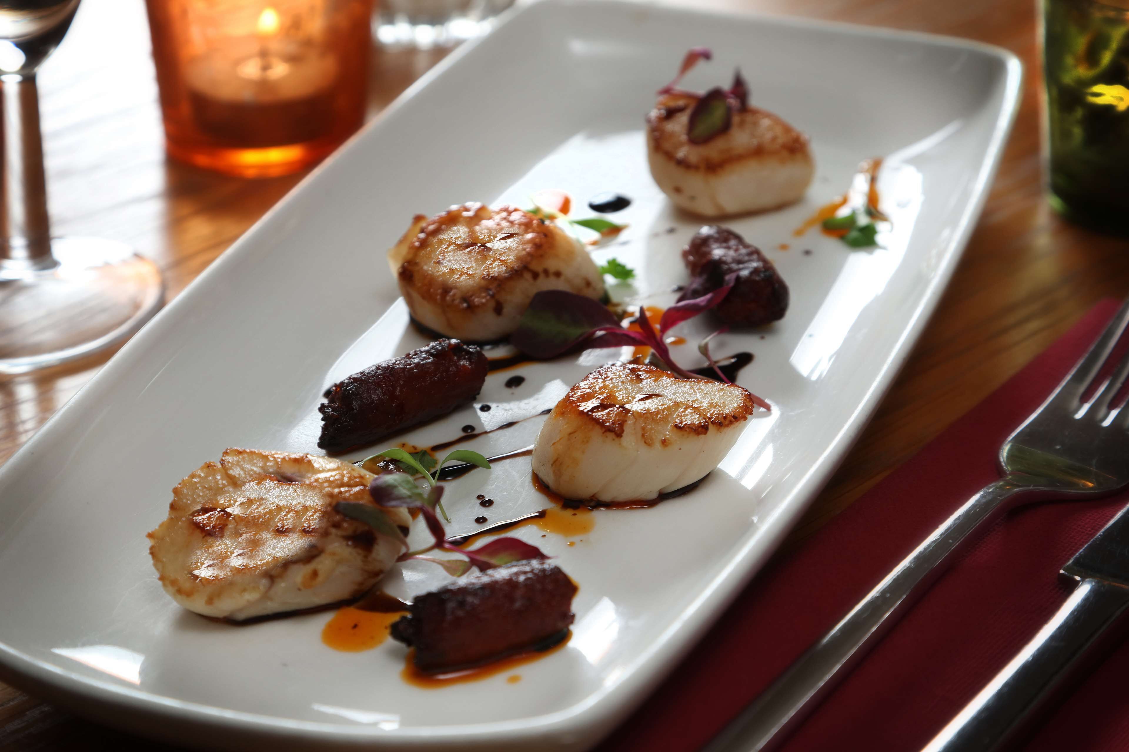 A rectangular white plate with seared scallops, a piece of meat, and some greens, accompanied by a glass of red wine and a candle in the background.