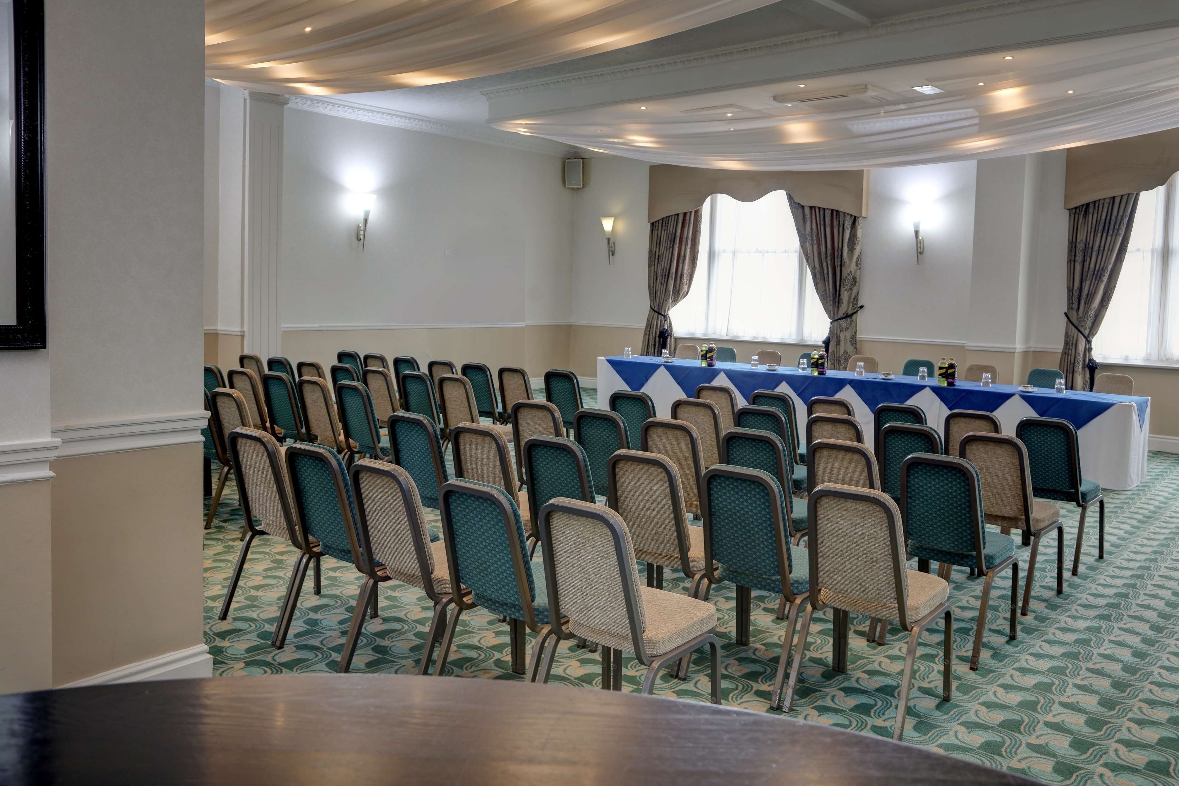 Un salón de conferencias con filas de sillas, una larga mesa con mantel azul y una alfombra estampada.