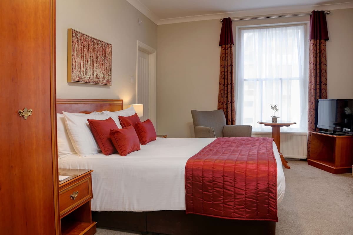 Ein Hotelzimmer mit einem großen Bett, einem Nachttisch, einem Sessel, einem kleinen Tisch mit Lampe, einem Fernseher und einem Fenster mit Vorhängen.