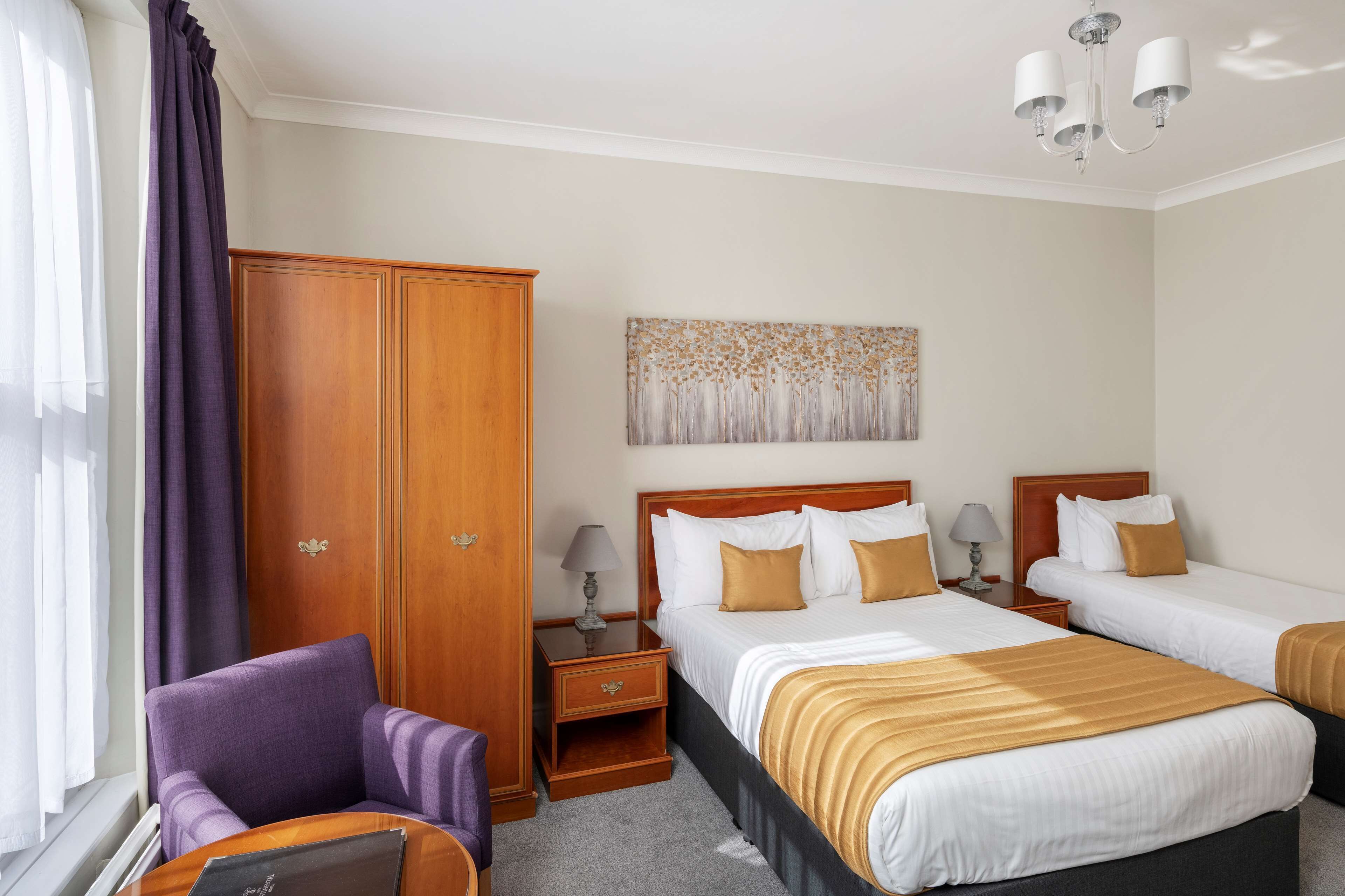 A hotel room with two beds, a wardrobe, a chair, a nightstand, and a painting on the wall.
