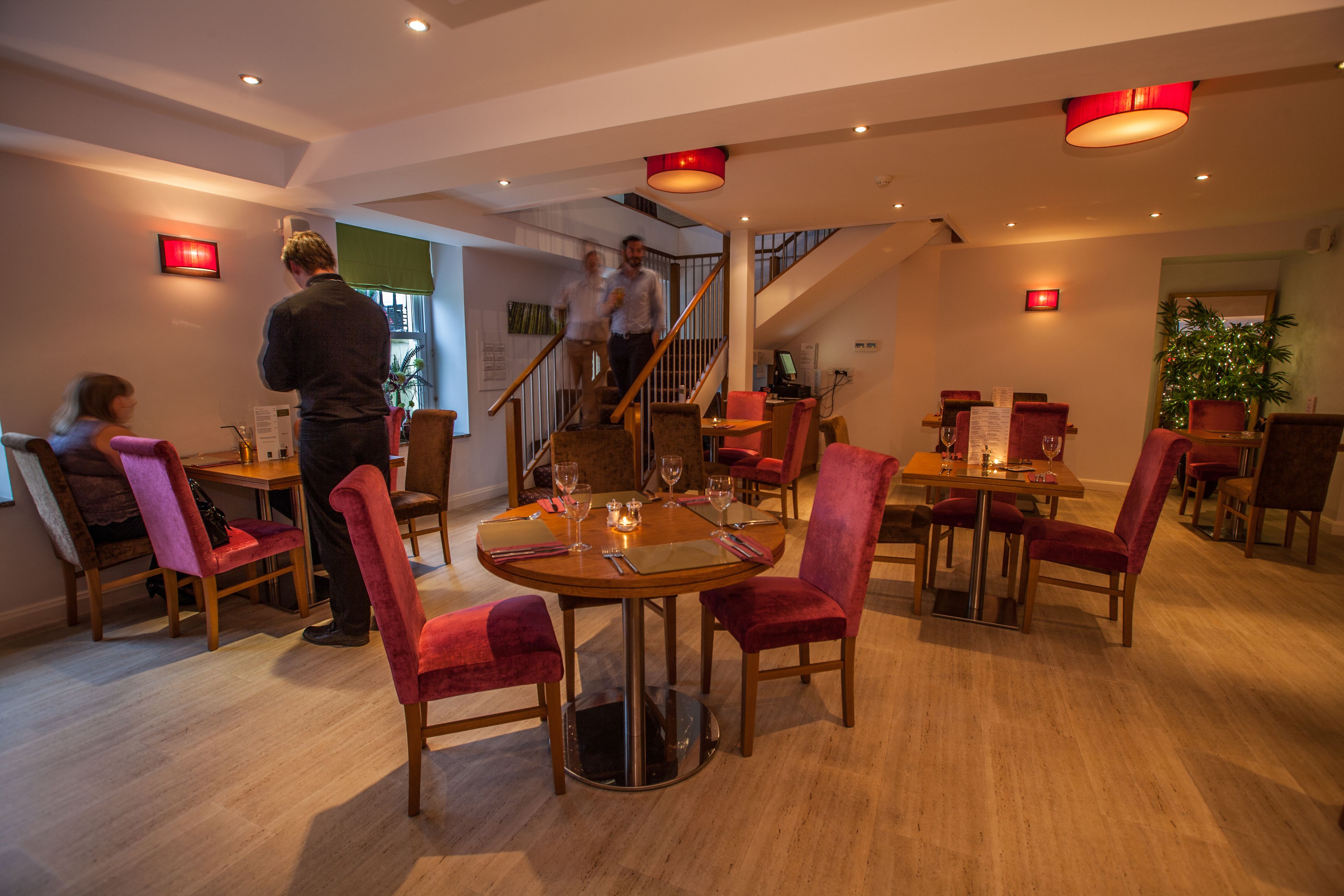 A dining area with round tables, red upholstered chairs, and a staircase in the background.