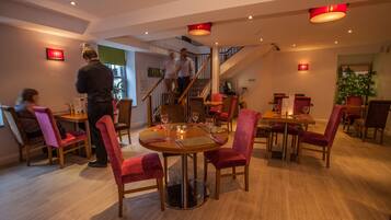 A dining area with round tables, red upholstered chairs, and a staircase in the background.