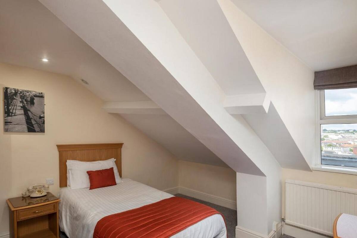Ein Schlafzimmer mit Schrägen, einem Bett mit roter Decke, einem Nachttisch mit Telefon und einem Fenster mit Blick auf Gebäude.