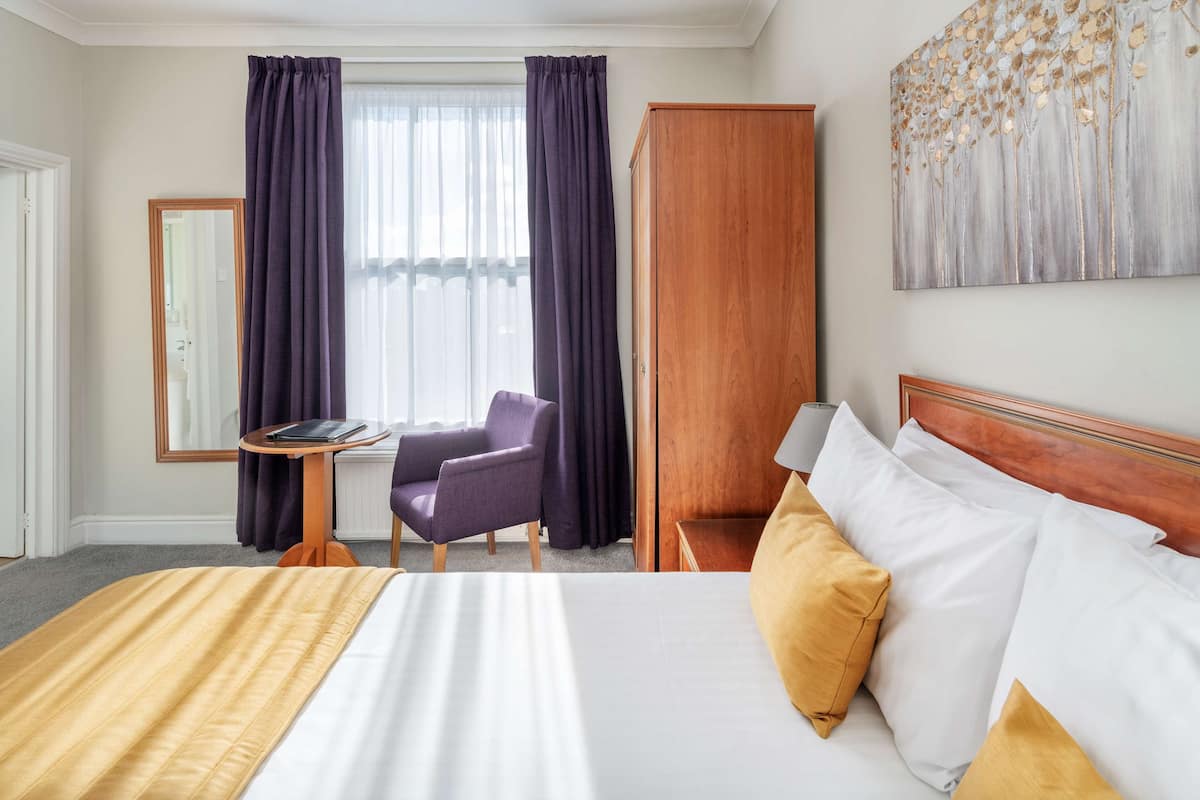 Ein Hotelzimmer mit Bett, Sessel, Schreibtisch, Kleiderschrank und einem Fenster mit Vorhängen.