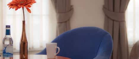 A room with a blue armchair, a round table with a white mug, a bottle, and a vase with a flower.