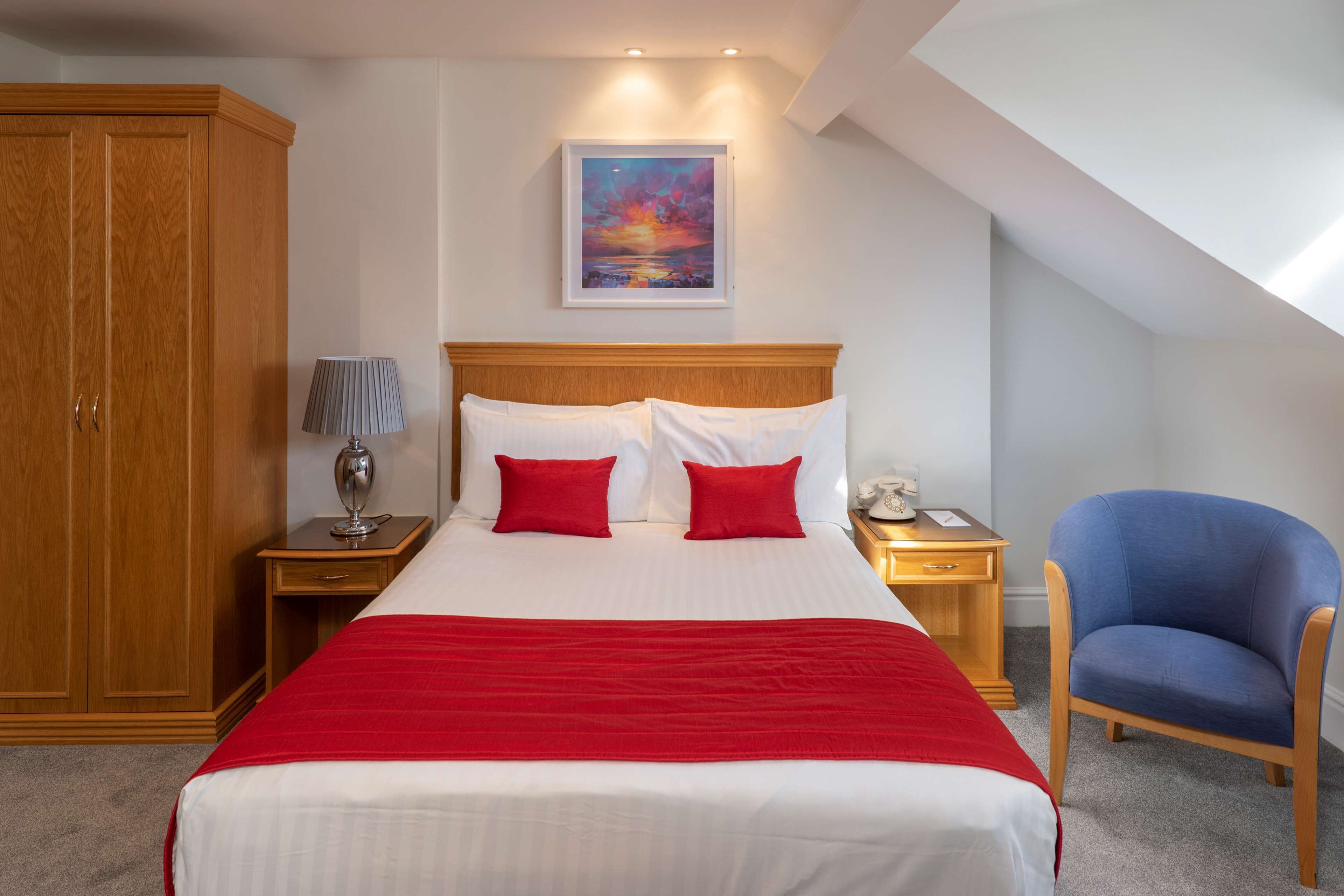 A hotel room with a large bed, a wooden wardrobe, a nightstand with a lamp, a chair, and a painting on the wall.