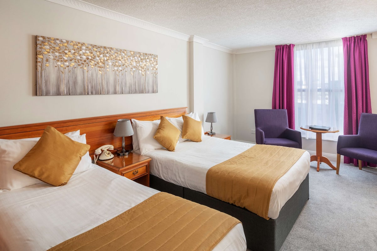 Ein Hotelzimmer mit zwei Betten, einem Bild an der Wand, einem Schreibtisch mit Lampe, violetten Sesseln und einem Fenster mit Vorhängen.