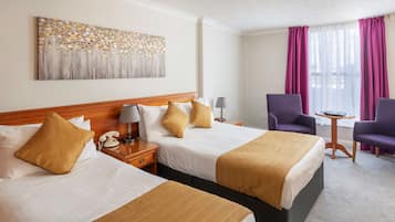 A hotel room with two beds, a painting on the wall, a desk with a lamp, purple chairs, and a window with curtains.