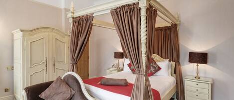 Un lit à baldaquin à quatre poteaux, avec une couvre-lit rouge et des rideaux marron, une commode blanche, une table de chevet avec une lampe, et un canapé marron avec des coussins.