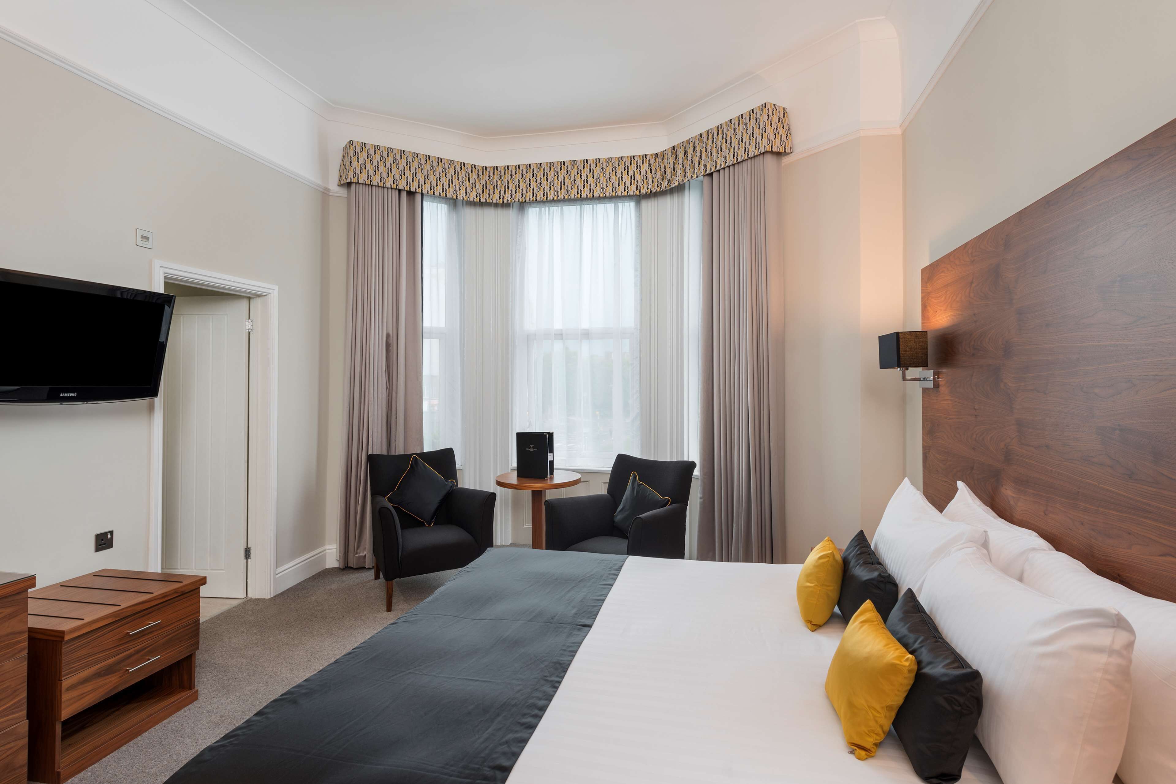 A hotel room with a large bed, two armchairs, a small table, a television, and a wooden headboard.