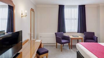 A hotel room with a bed, a desk with a chair, a TV, a mirror, and two windows with curtains.