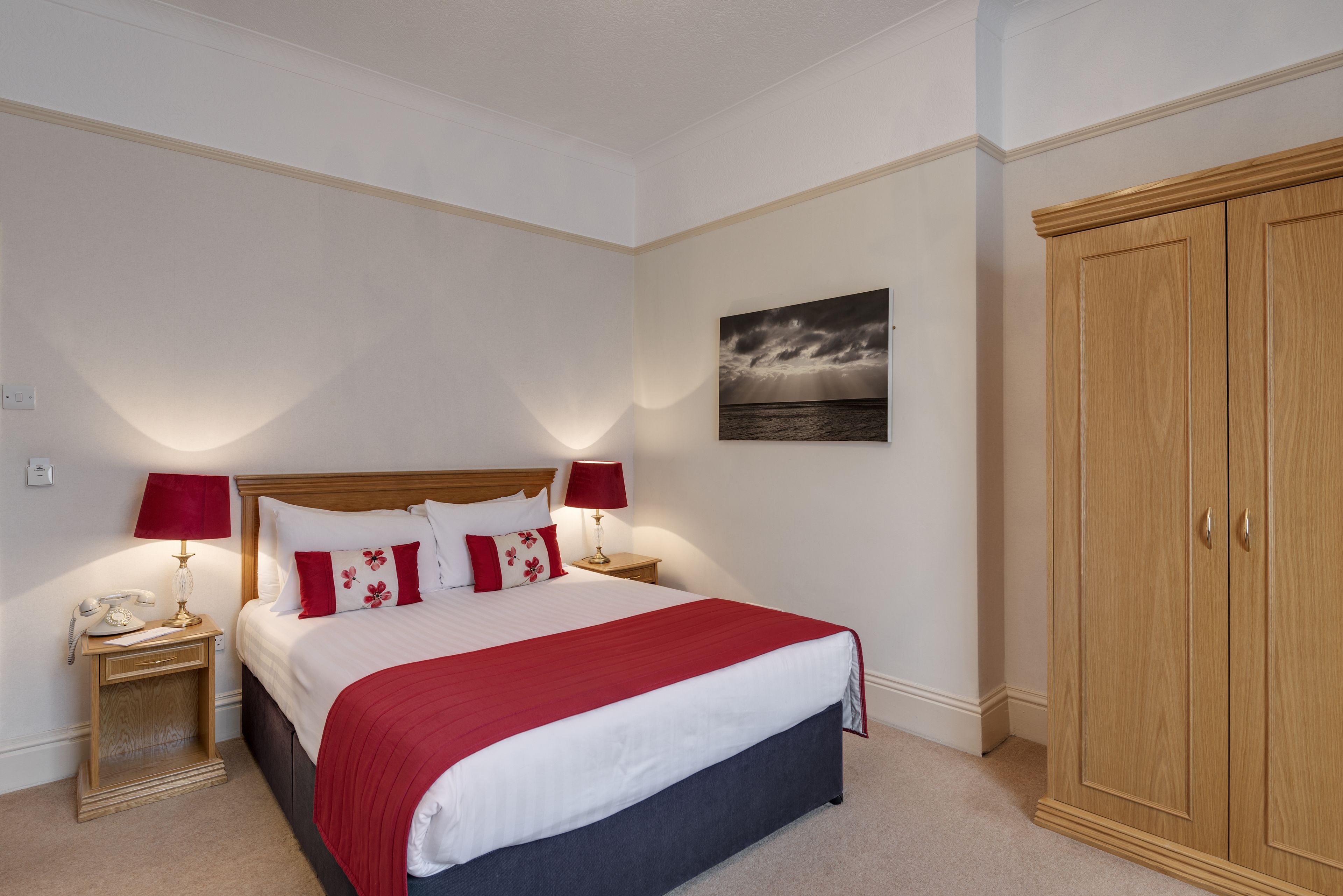 A neatly arranged bedroom with a bed, bedside lamps, a nightstand, a wardrobe, and a wall-mounted picture.