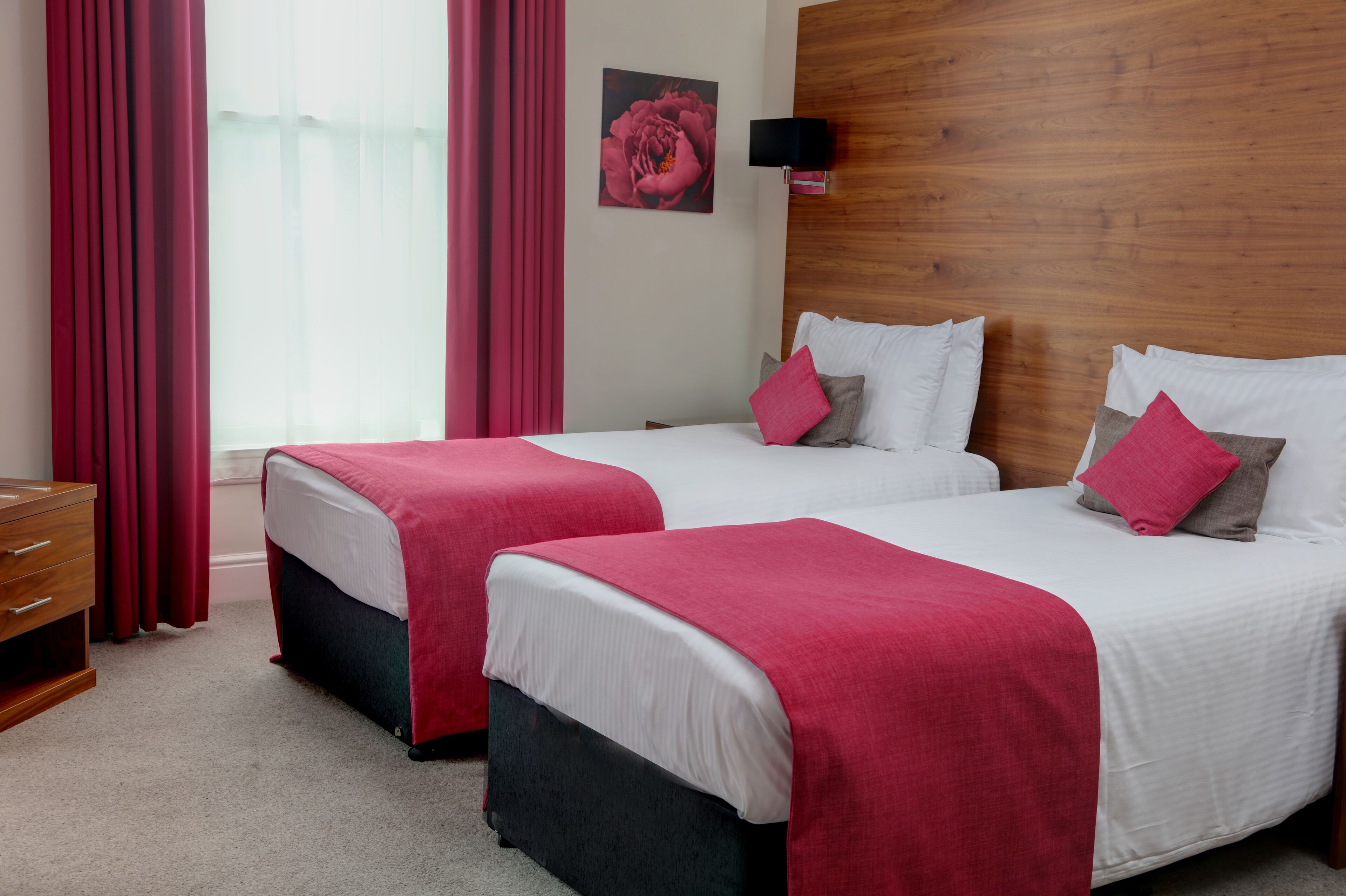 Une chambre d’hôtel avec deux lits, chacun recouvert d’une courtepointe blanche et rouge, dotée d’une tête de lit en bois, d’une commode en bois et d’un rideau rouge.