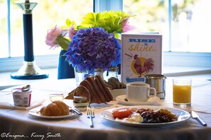 A breakfast table with croissants, a plate of tomatoes, a cup of coffee, a glass of orange juice, and a vase with flowers.