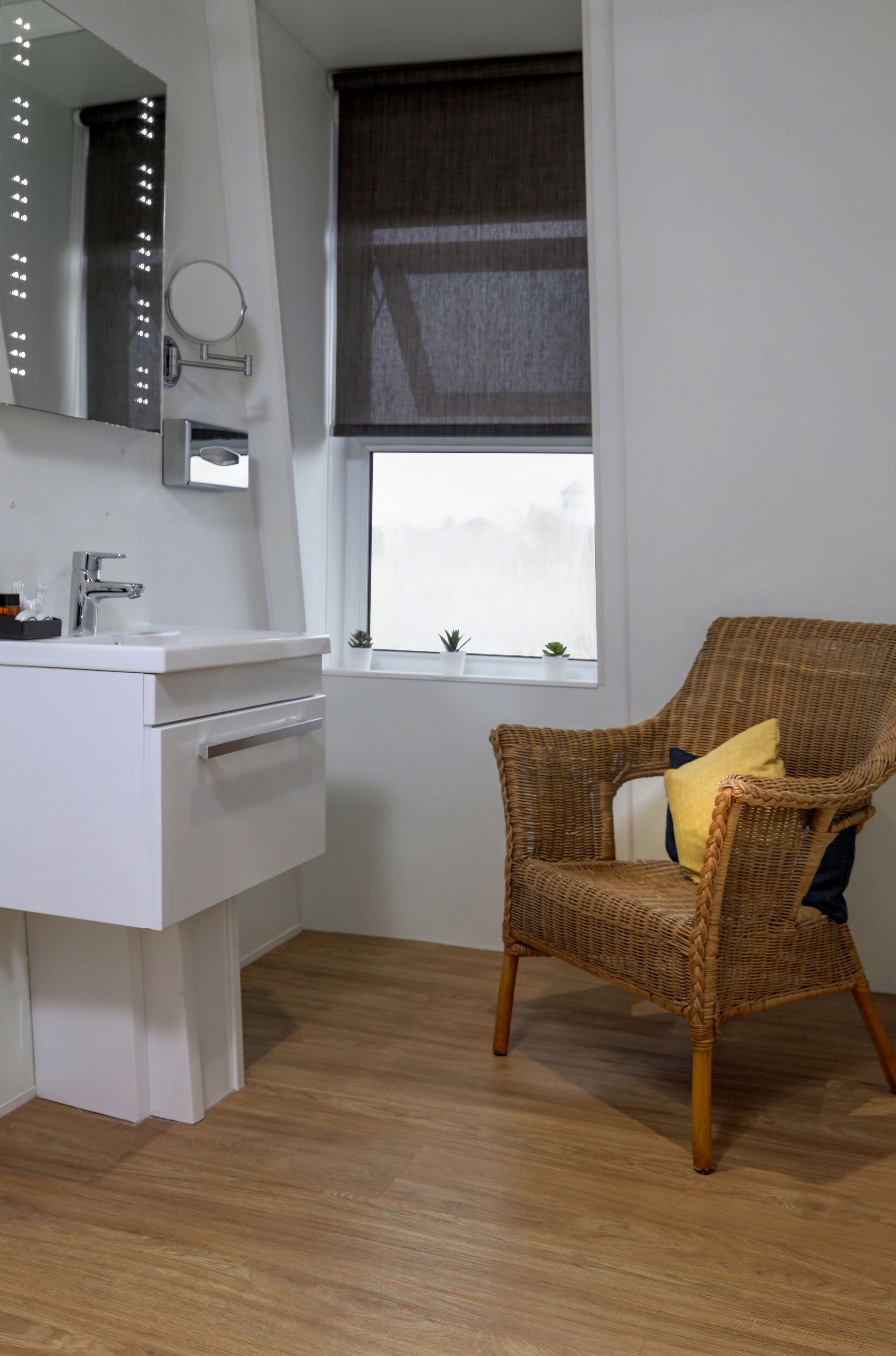 A bathroom with a white vanity, a mirror with lights, a wicker chair with a yellow pillow, and a window with a view.