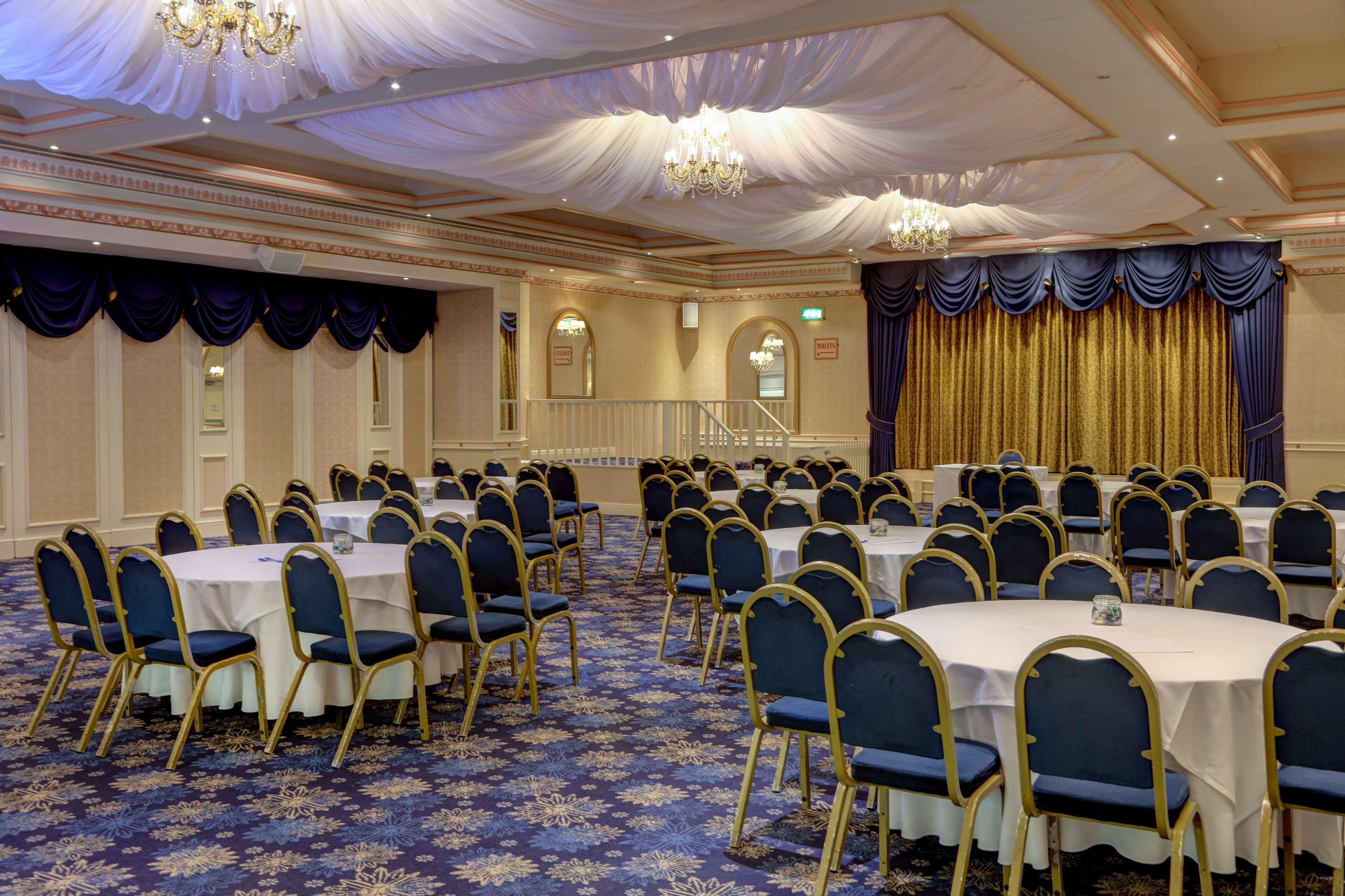 A banquet hall with round tables, blue chairs, and a gold curtain backdrop.
